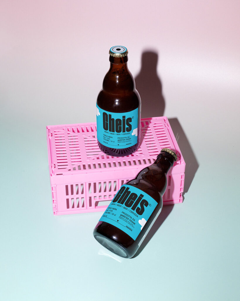 Progetto di fotografia commerciale di METWISH Studio Creativo Multidisciplinare, Firenze e Lucca di due bottiglie di birra Cheis Triple NEIPA con etichetta azzurra, appoggiate su una cassetta rosa da trasporto.